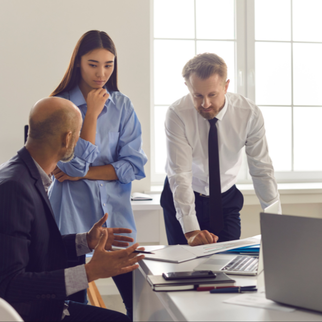Trois professionnels discutent autour d'une table, avec un homme au crâne rasé, une femme avec les bras croisés, et un homme en chemise blanche, concentrés sur un document dans un bureau.