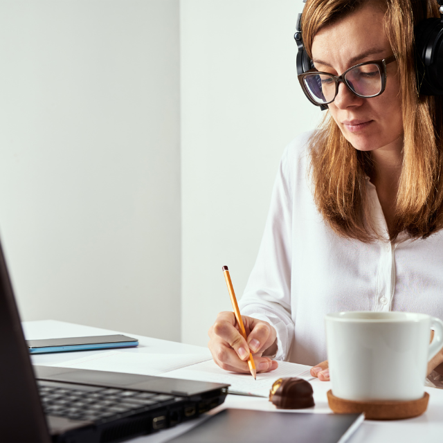 Personne assise à un bureau, portant des écouteurs et des lunettes, écrivant sur une feuille de papier, avec un ordinateur portable et une tasse, dans une atmosphère concentrée et studieuse.